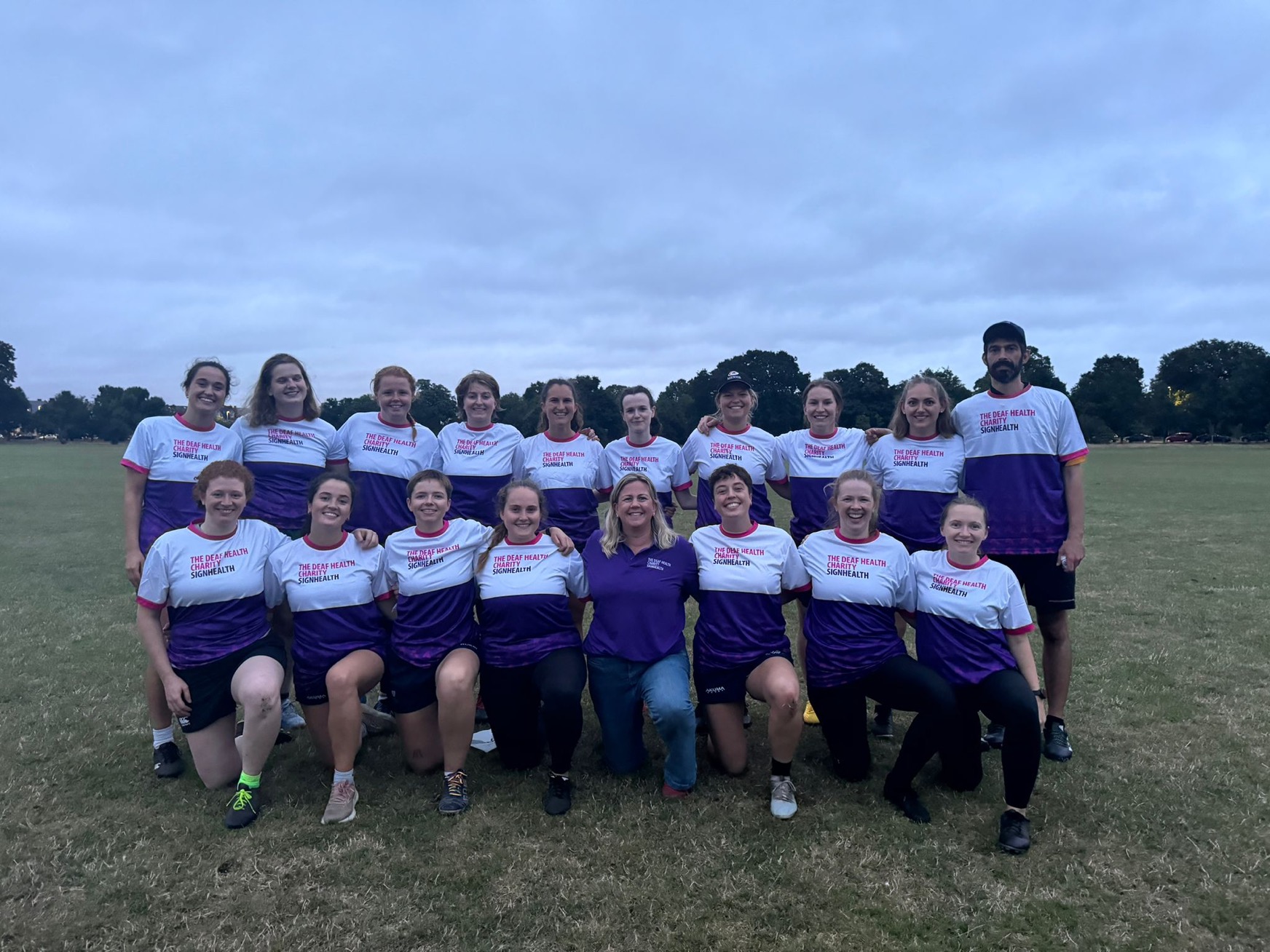 Group of rugby players in their kit and fundraising SignHealth t-shirts with fundraising officer all posing in a team photo on a field.