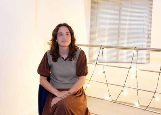 Young person, female presenting with long brown hair, brown dress sat with fairy lights in background.