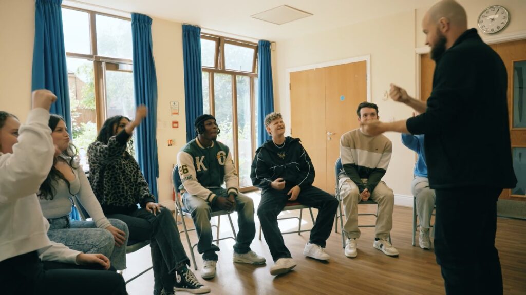 6 young people sitting in a semi-circle, some are raising their hands, and a man is standing in front of them giving a presentation