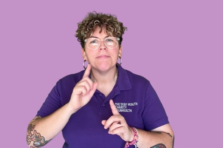 plain purple background with BSL user white female with short dark curls wearing the SignHealth brand t shirt