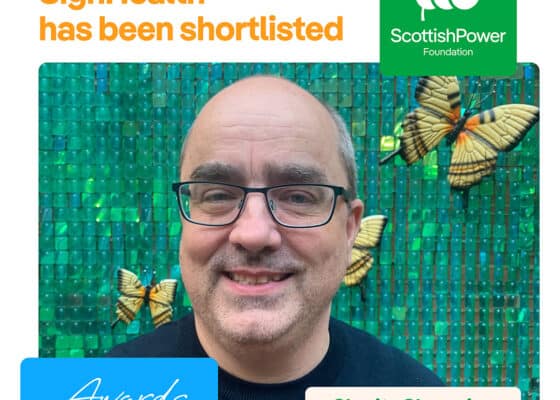 white male with a bald head, wearing glasses and a dark top aganist a green tiled wall with butterflies. Green graphic logo for scottish power awards.